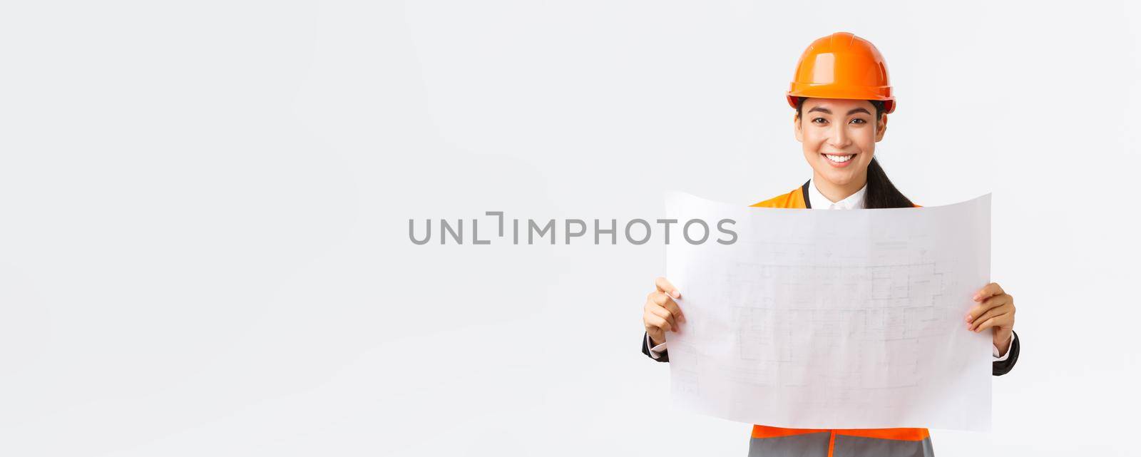 Excited smiling asian female engineer at construction area study blueprints, looking pleased while reading documents, building plan, standing in safety helmet over white background.