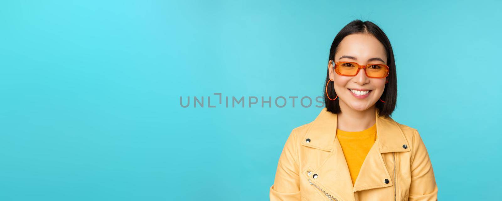 Close up portrait of smiling stylish asian woman in sunglasses, looking happy at camera, posing in trendy yellow jacket, blue background by Benzoix