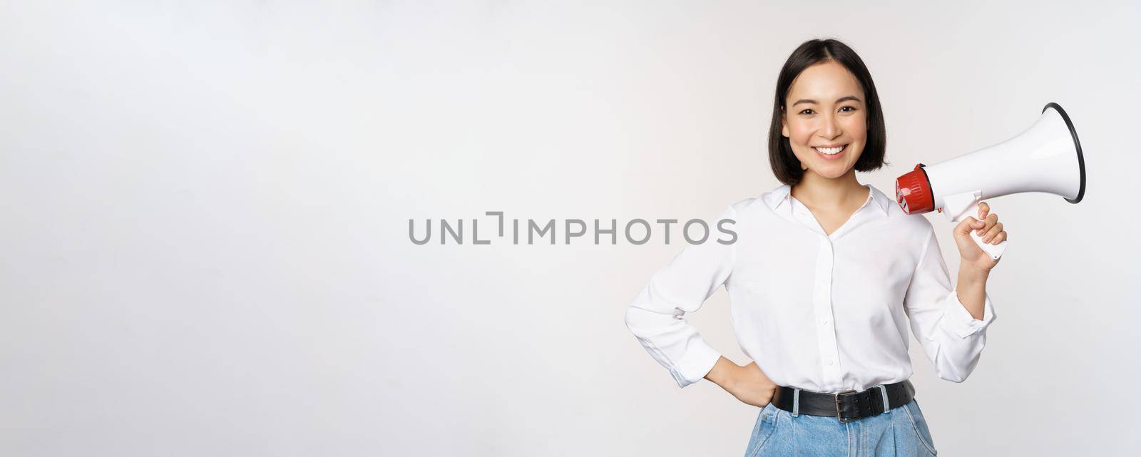 Smiling young asian woman posing with megaphone, concept of news, announcement and information, standing over white background by Benzoix