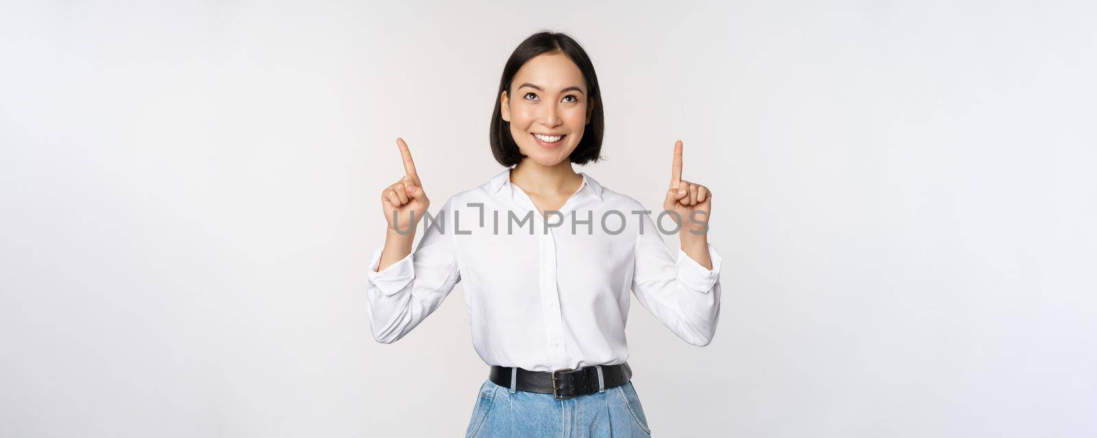 Enthusiastic asian business woman pointing, looking up with happy smiling face, showing company logo or banner, standing over white background by Benzoix