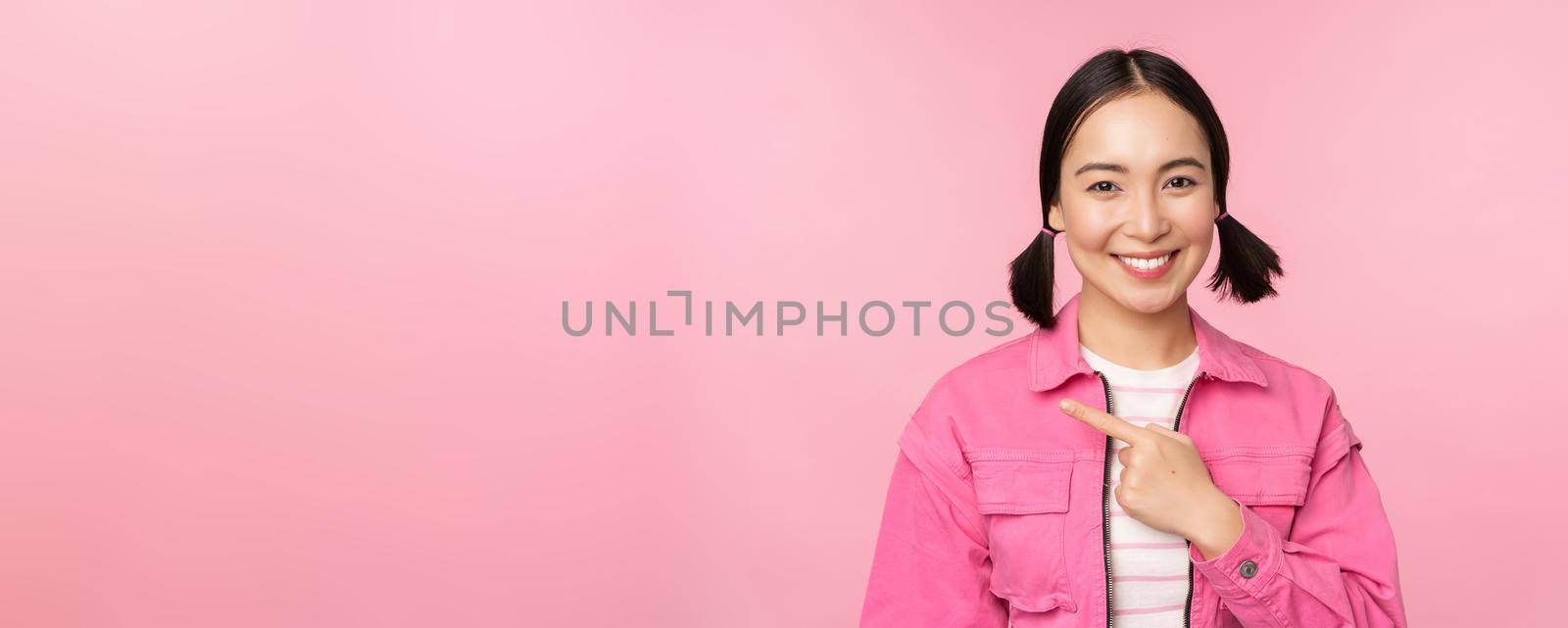 Portrait of beautiful smiling asian girl, pointing finger left, showing advertisement, banner promo, standing over pink background by Benzoix