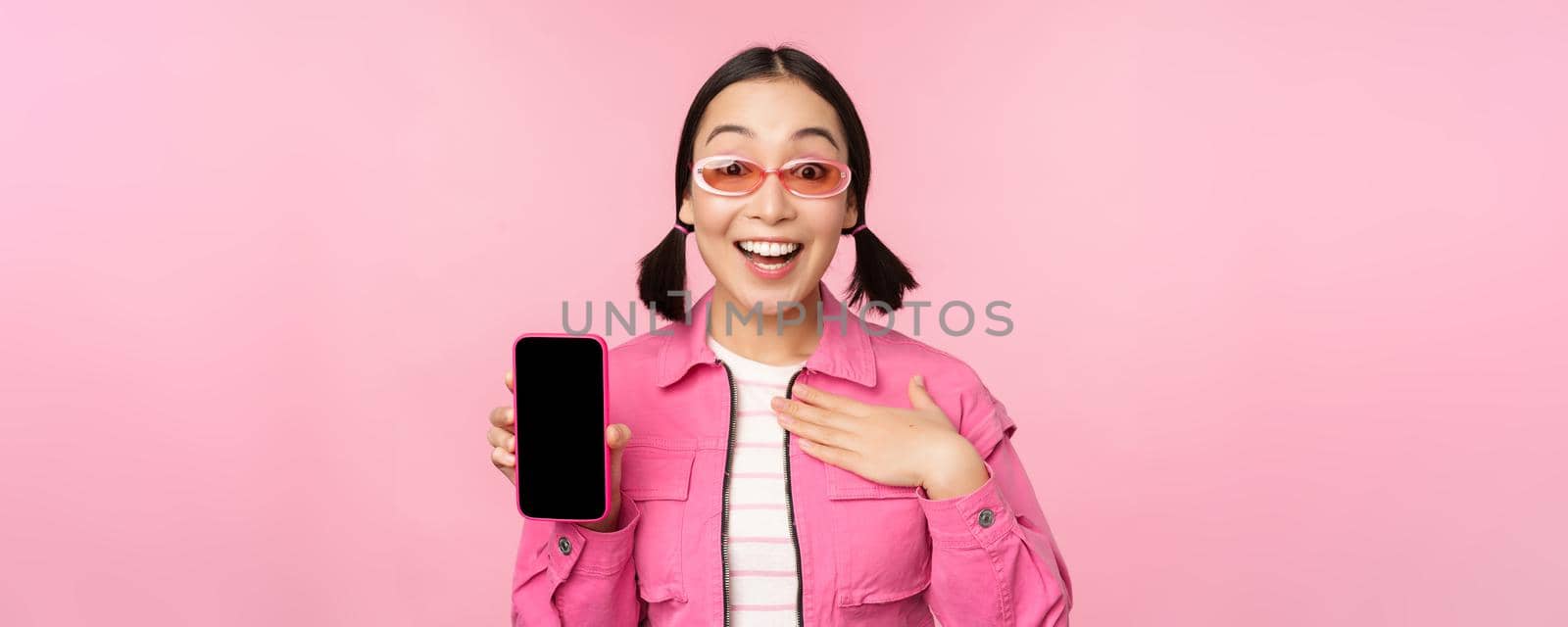 Image of surprised girl showing mobile phone app screen, smartphone display, application interface, standing against pink background by Benzoix