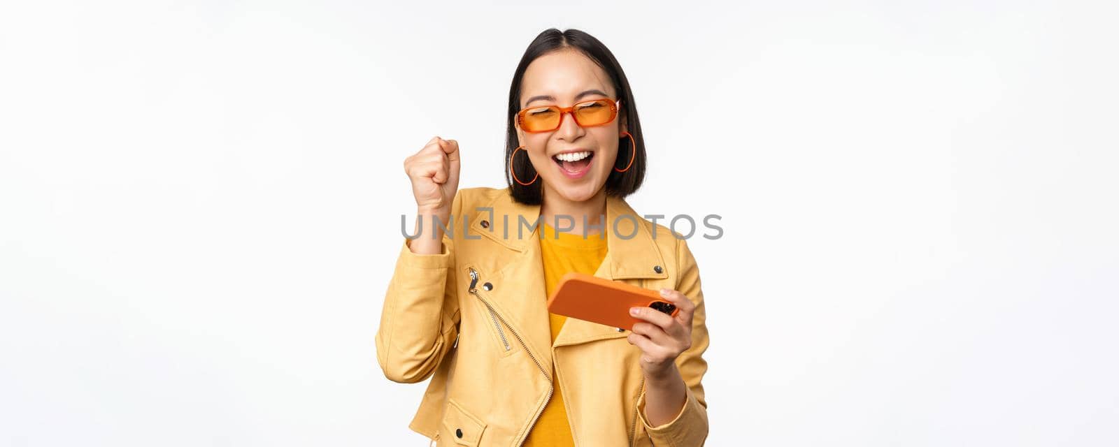 Image of stylish korean girl dancing with smartphone, laughing happy and smiling, standing over white background. Copy space