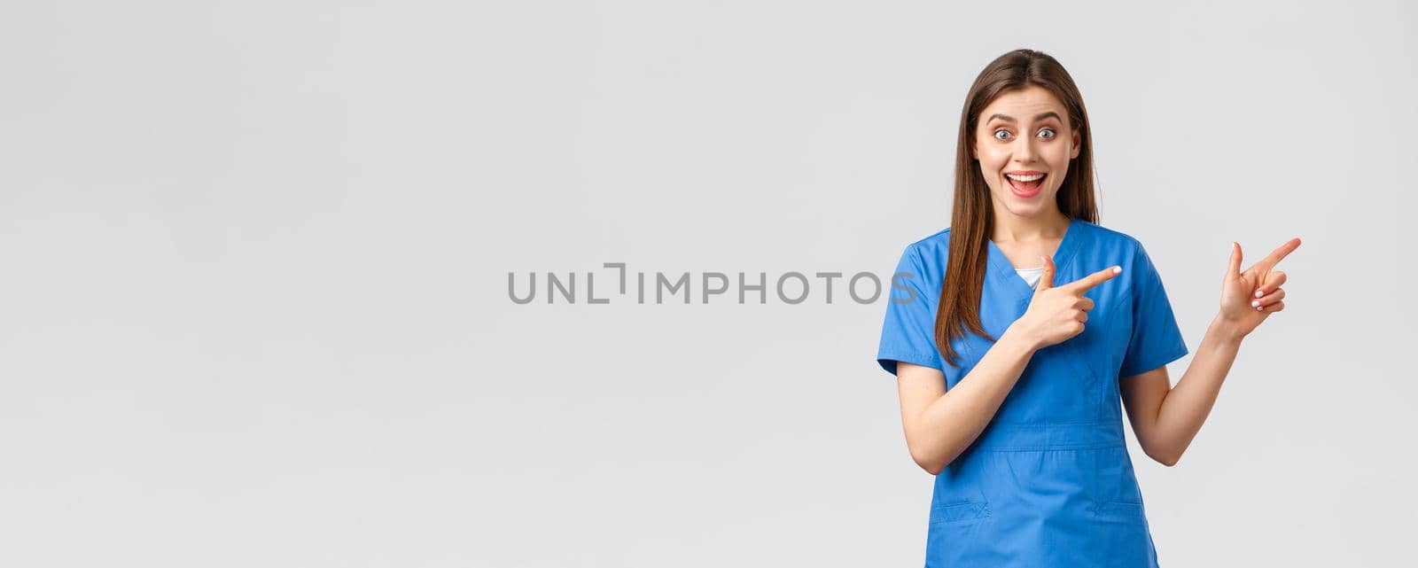 Healthcare workers, prevent virus, insurance and medicine concept. Excited happy female nurse or doctor in blue scrubs pointing fingers right, look amused, inform about promo or good news.