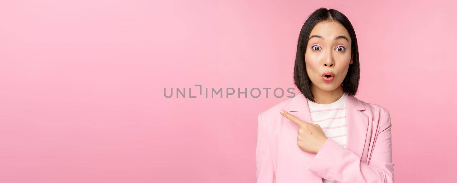 Portrait of businesswoman with surprised face, pointing finger left, showing smth interesting, standing over pink background in office suit.