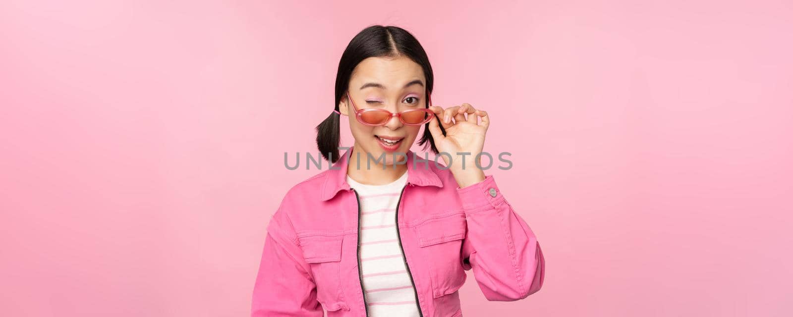 Eyewear advertisement. Stylish modern asian girl touches sunglasses, wears pink, poses against studio background. Copy space.