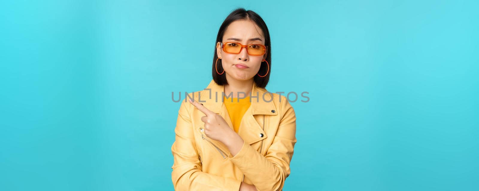 Portrait of disappointed korean woman in sunglasses, stylish girl points finger left and grimacing displeased, dislike smth, stands over blue background.