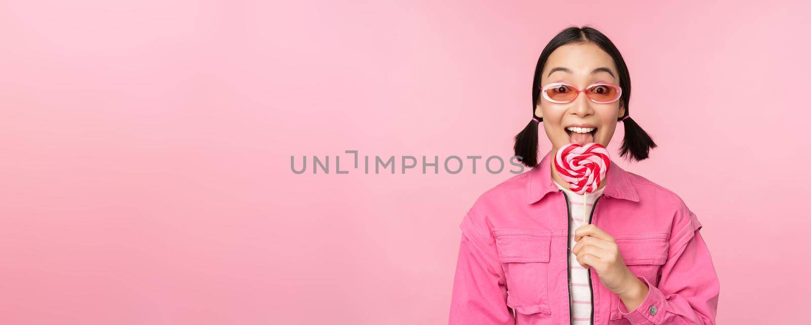 Stylish korean girl licking lolipop, eating candy and smiling, standing in sunglasses against pink background by Benzoix