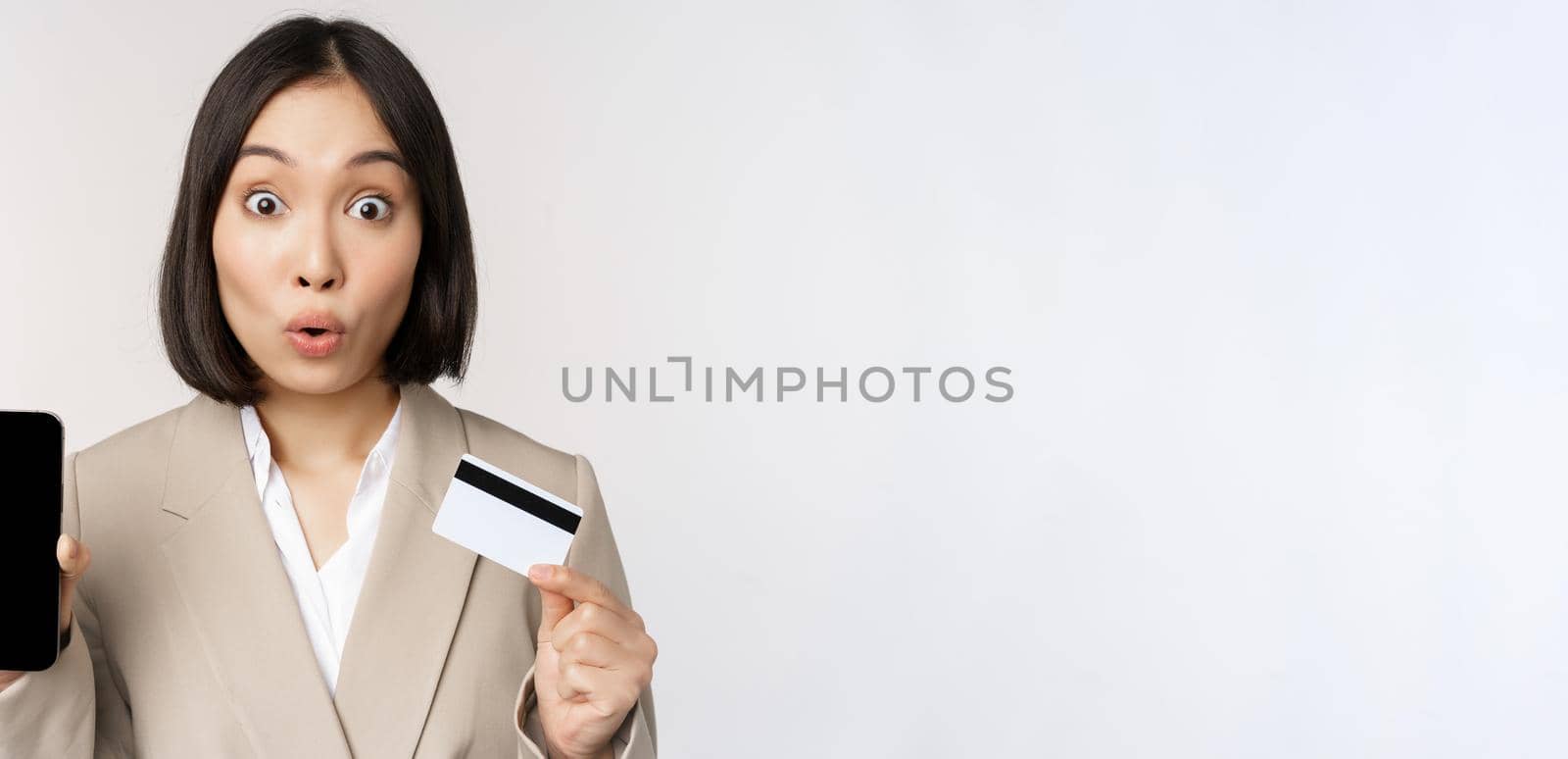 Corporate woman with happy, enthusiastic face, showing credit card and smartphone app screen, standing in suit over white background.