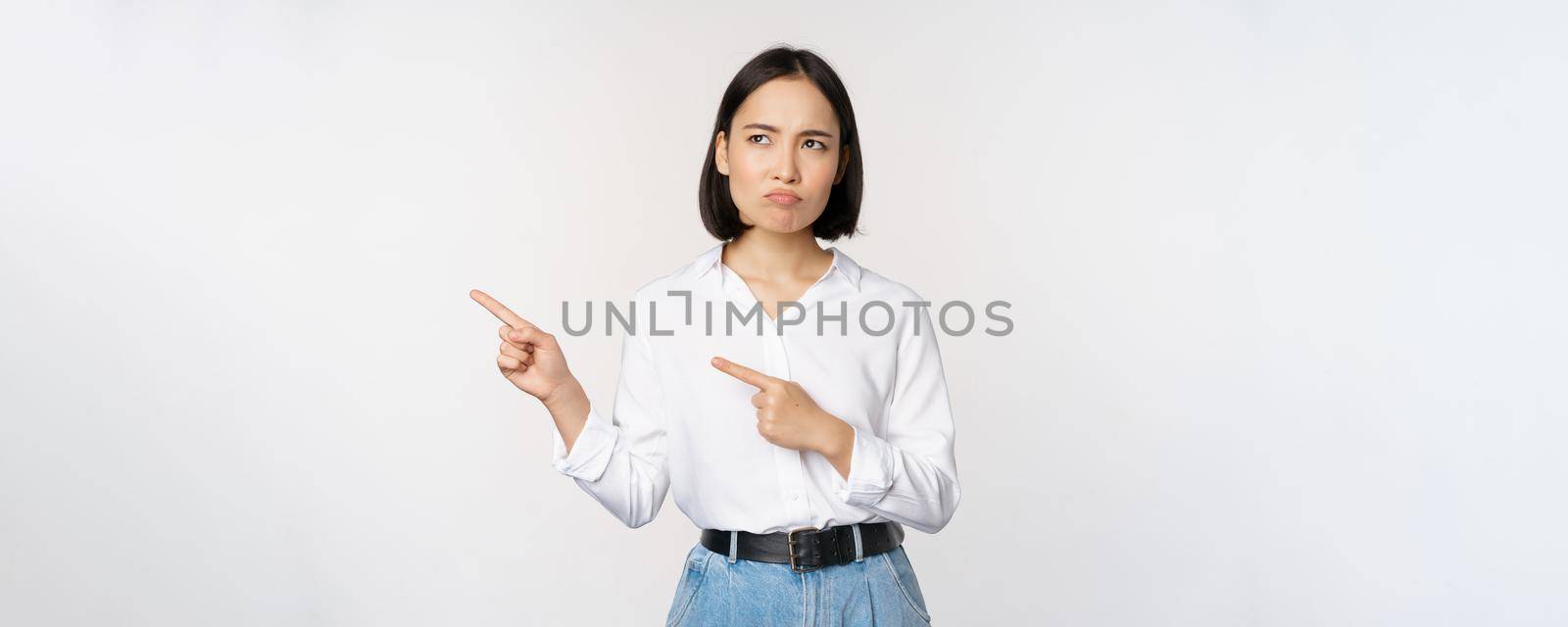 Disappointed, sad young asian woman pointing and looking left with upset, sulking face expression, standing over white background by Benzoix