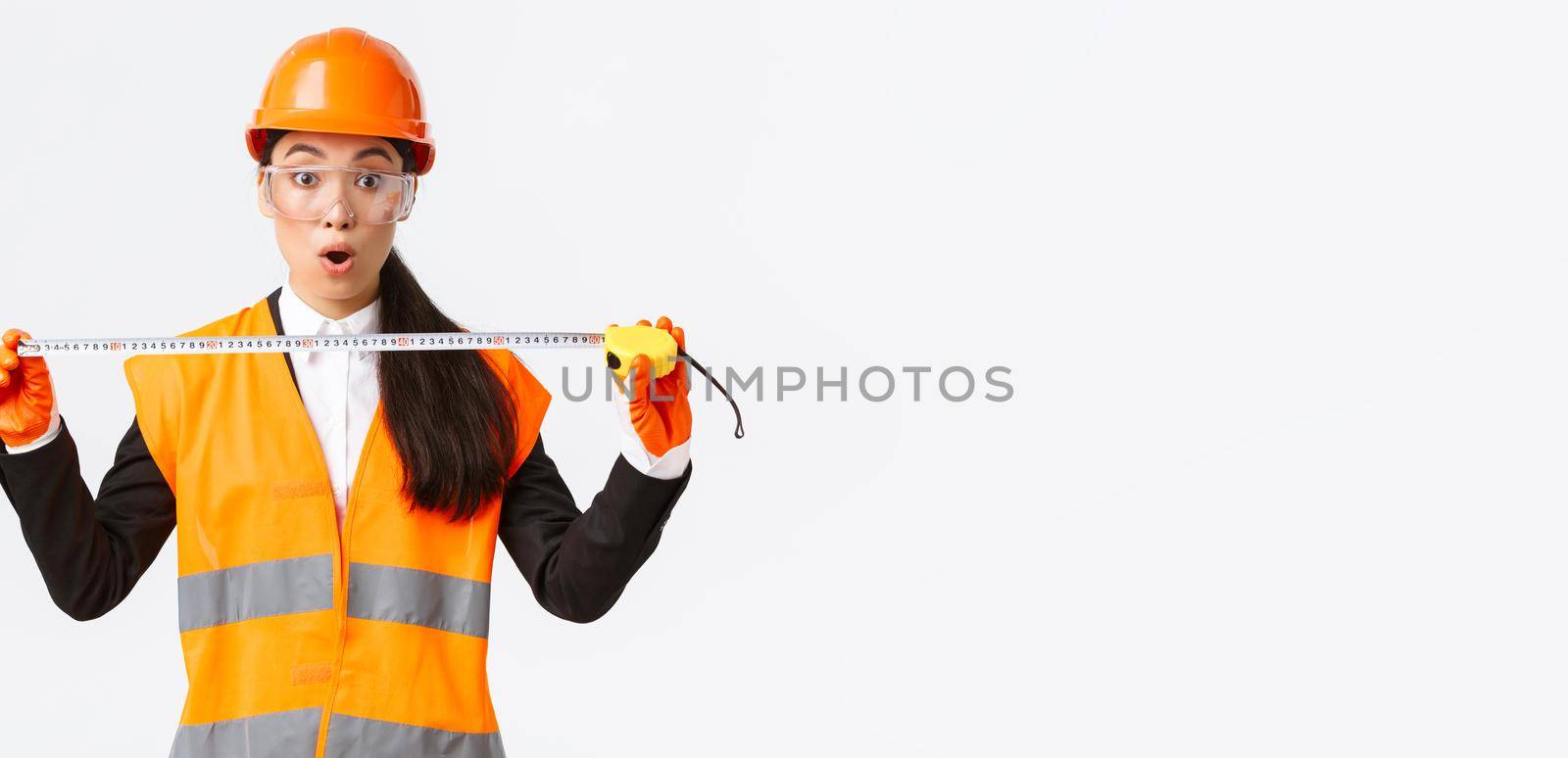 Surprised and excited asian female construction engineer, industrial worker in safety helmet and uniform, measuring layout, holding tape measure and stare amazed at camera, white background by Benzoix