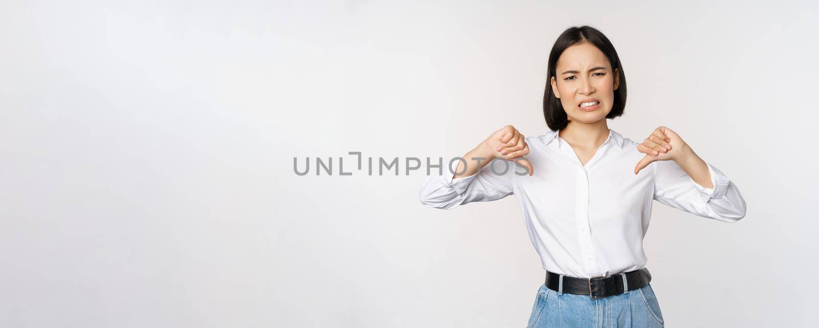 Dislike. Disgusted asian woman showing thumbs down and grimacing, commenting on smth bad, disagree, complaining, standing over white background.