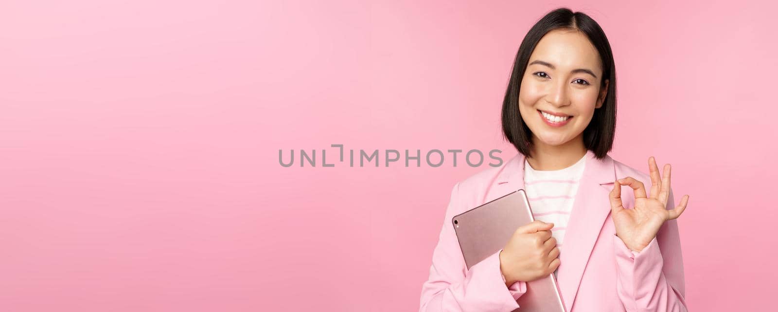 Portrait of corporate woman, girl in office in business suit, holding digital tablet, showing okay, recommending company, standing over pink background.