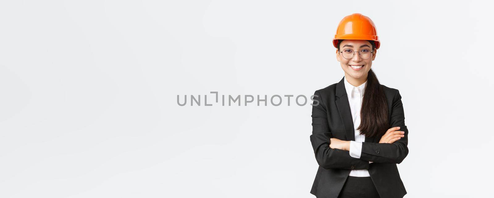 Professional smiling asian businesswoman in safety helmet and suit cross arms and looking confident, selling new buildings. Construction manager, engineer showing around, greeting investors.