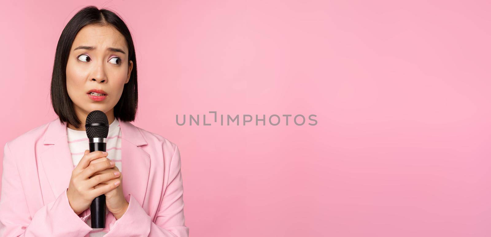 Insecure asian businesswoman giving speech, scared of talking in public, using microphone, standing in suit over pink background by Benzoix