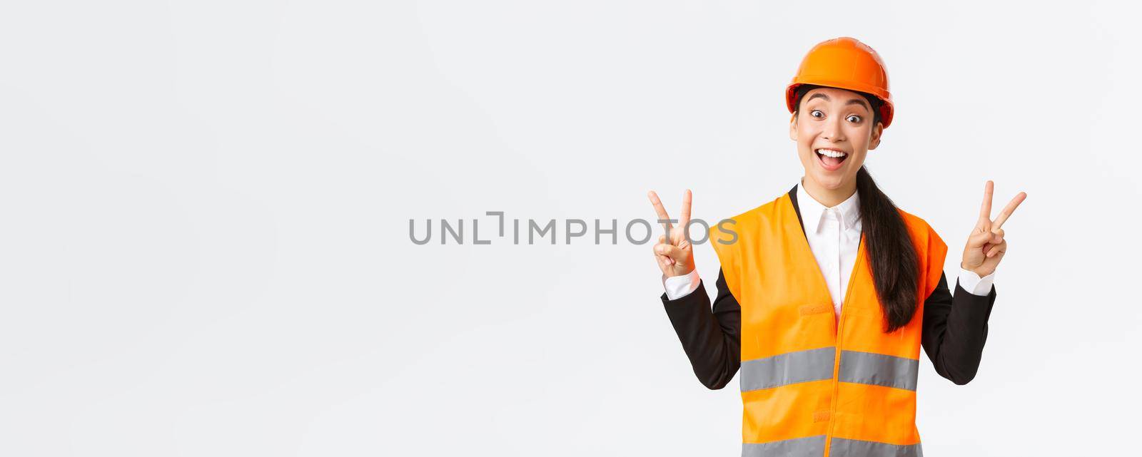 Happy smiling asian female engineer, architect in safety clothins and helmet, showing peace kawaii sign and looking upbeat, company winning tender on construction and building, white background.
