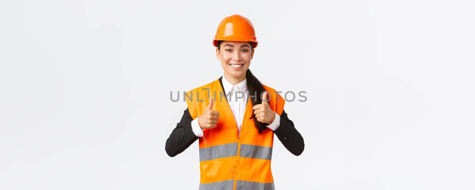 Upbeat pleased asian female architect giving permission, proud with result, standing in safety helmet and reflective jacket, showing thumbs-up in approval, guarantee best construction quality.