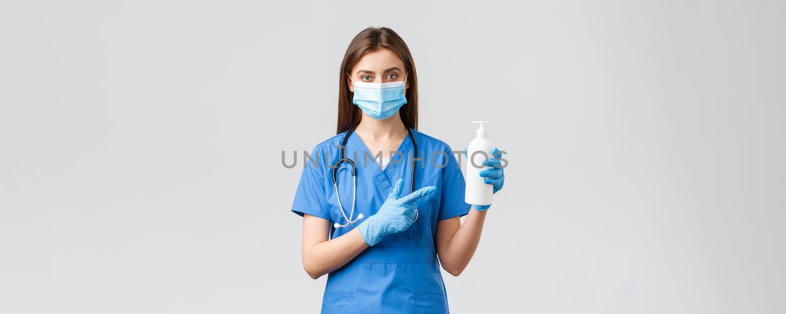 Covid-19, preventing virus, health, healthcare workers and quarantine concept. Young pretty female nurse or doctor in blue scrubs, medical mask and gloves, pointing at hand sanitizer, soap.