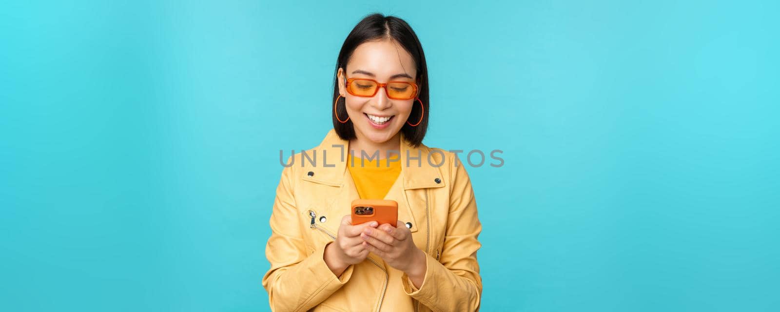 Smiling asian girl in sunglasses, using smartphone app, holding mobile phone, standing over blue background by Benzoix