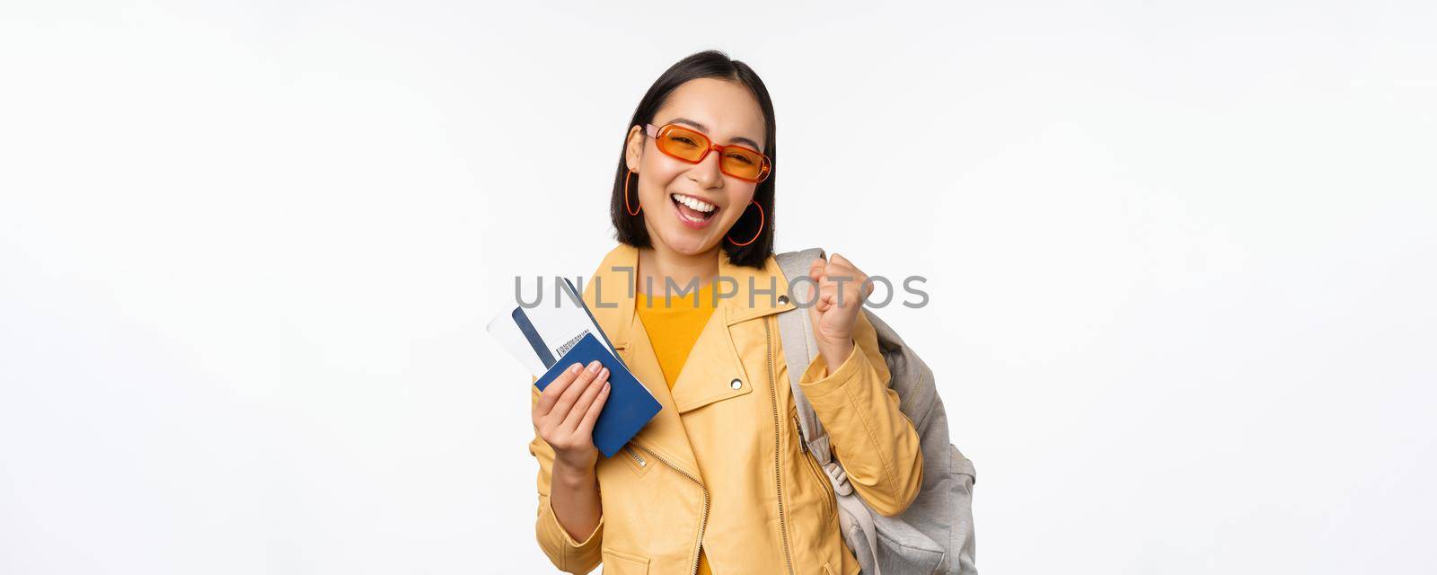 Happy asian girl going on vacation, holding passport and flight tickets, backpack on shoulder. Young woman tourist travelling abroad, standing over white background by Benzoix