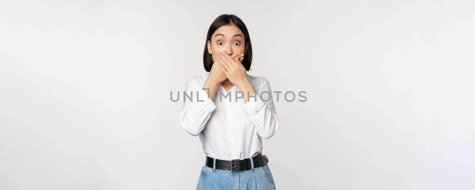 Shocked young asian girl close mouth, cover lips with hands and looking amazed, standing over white background. Taboo gesture