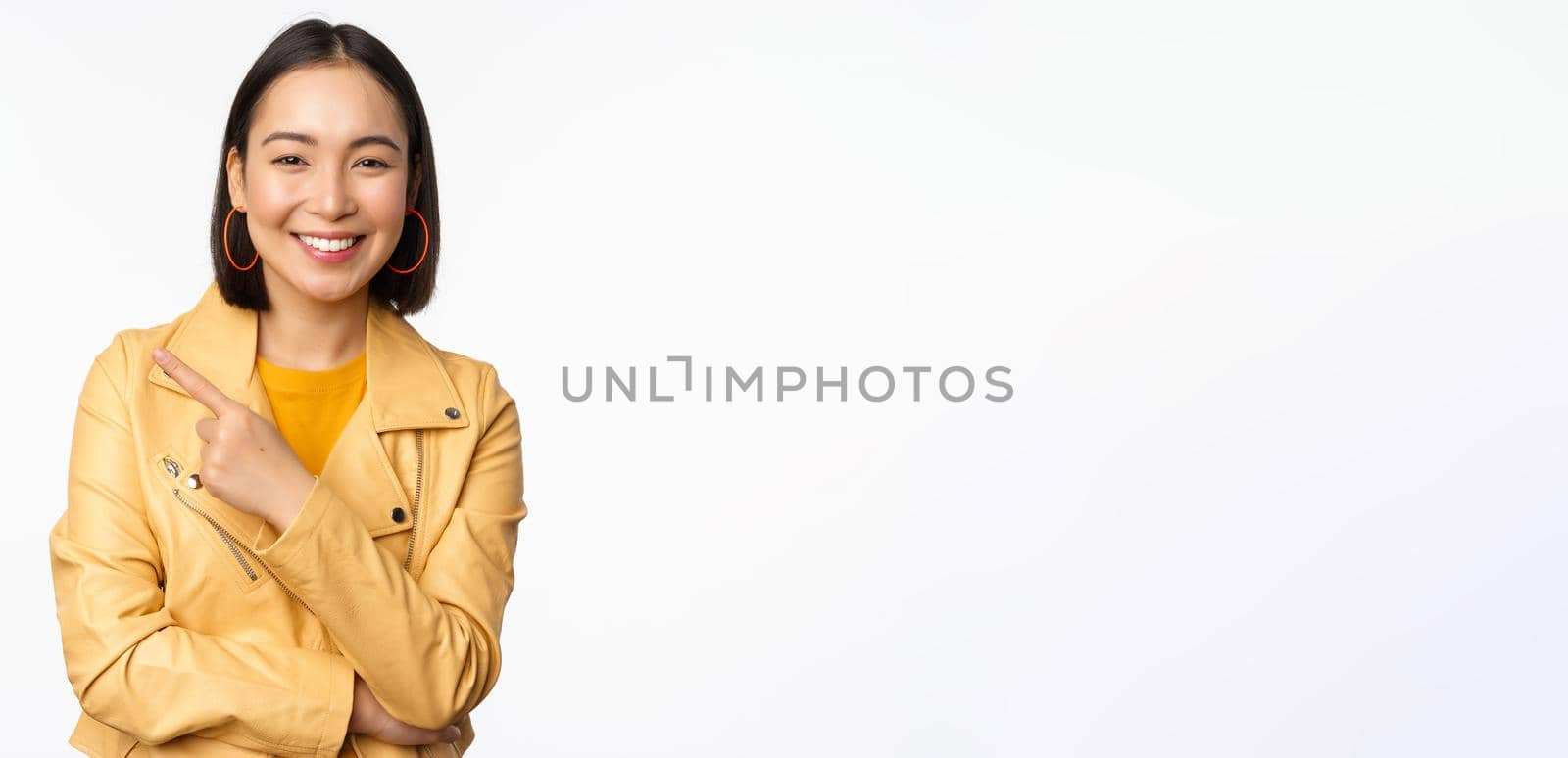 Portrait of smiling asian girl pointing finger left, showing logo or copy space, advertisement on empty side, standing over white background by Benzoix