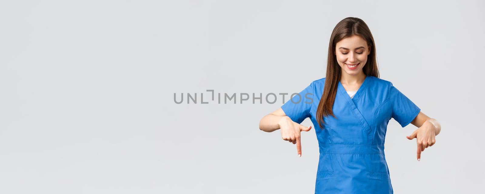 Healthcare workers, prevent virus, covid-19 test screening, medicine concept. Happy and pleased attractive female nurse or doctor, physician in blue scrubs, pointing looking down at advertisement.