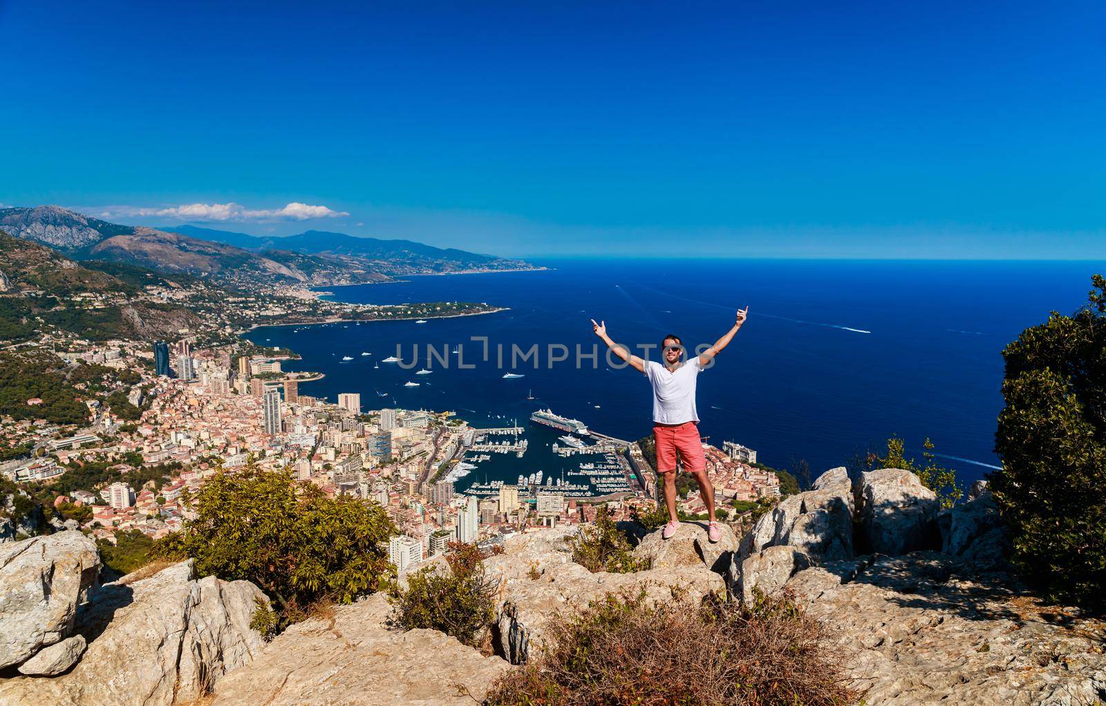 A handsome young man stands and jubilantly with his hands raised on a mountain with the name Of a Head of dog, the Principality of Monaco in the background in clear sunny weather. High quality photo