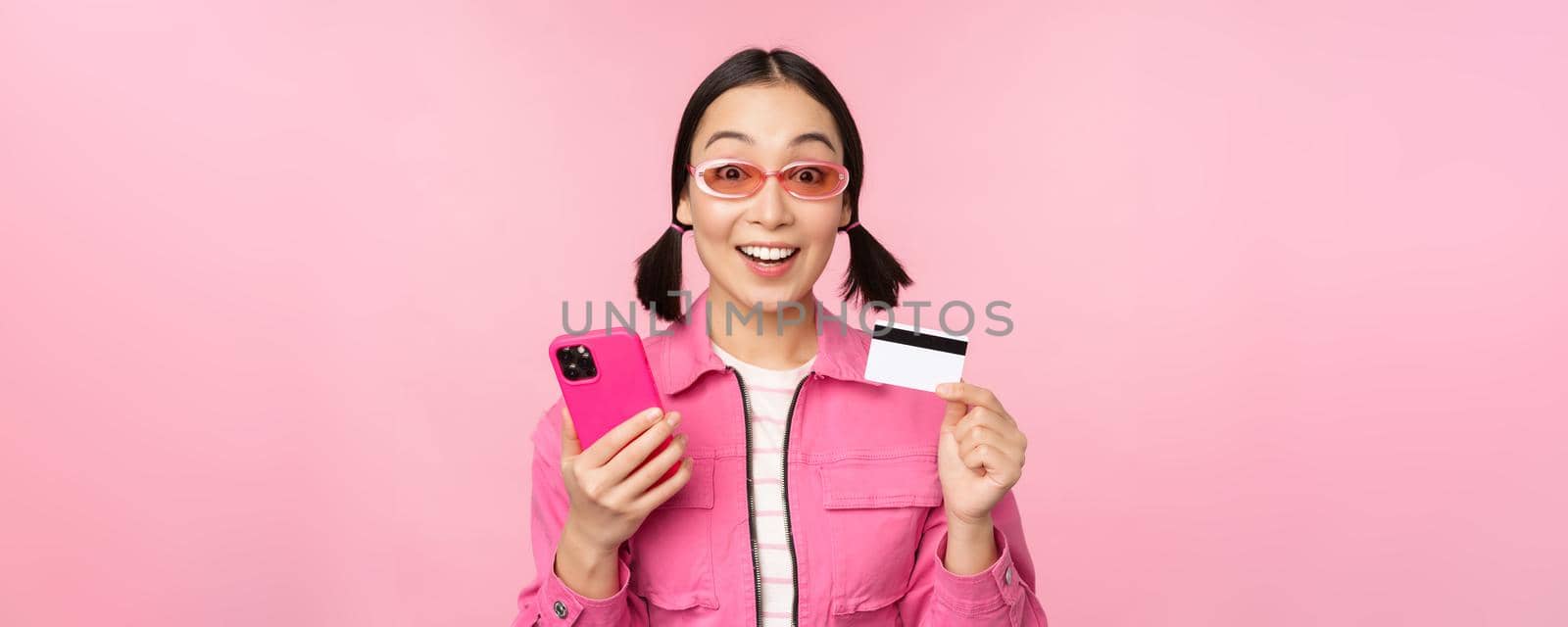 Online shopping. Smiling asian girl shopper, holding smartphone and credit card, paying in mobile app, standing over pink background by Benzoix