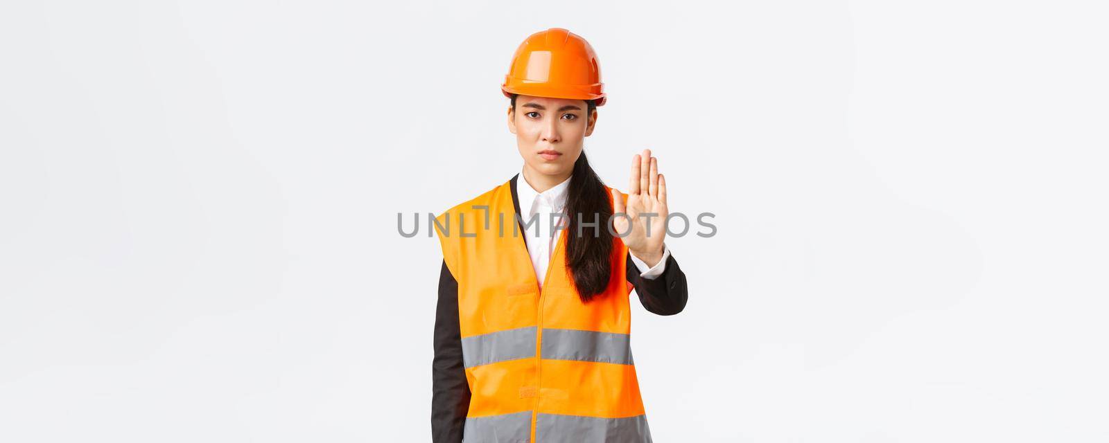 Serious-looking disappointed asian female architect, construction manager at working area wearing safety helmet, showing stop gesture, prohibit action, forbid trespassing, white background.