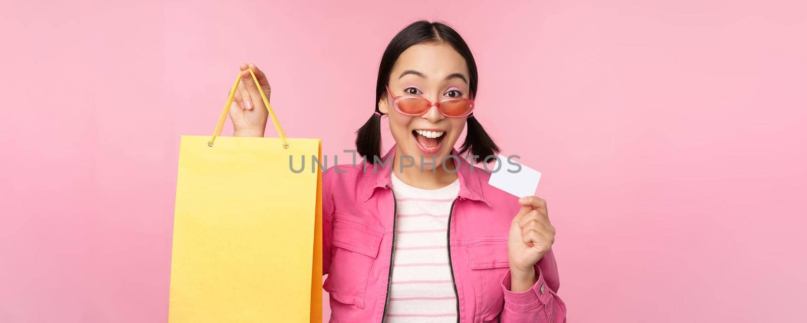 Happy young asian woman shows credit card and shopping bag, store sale announcement, buying smth in shop, posing against pink background.