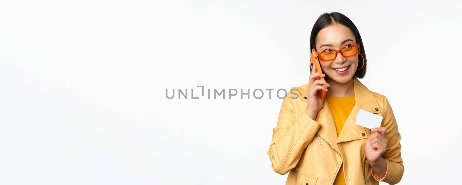 Stylish asian female model, talking on smartphone and showing credit card, standing over white background. Copy space