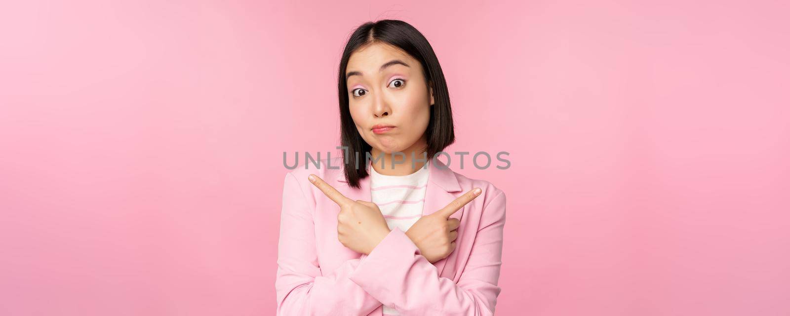 Decision concept. Young asian businesswoman, corporate worker pointing fingers sidways, two directions, showing variants, looking indecisive while choosing, pink background by Benzoix