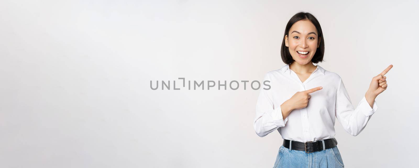 Portrait of beautiful asian woman, saleswoman pointing fingers right and showing info, banner or logo, sale advertisement, standing over white background.