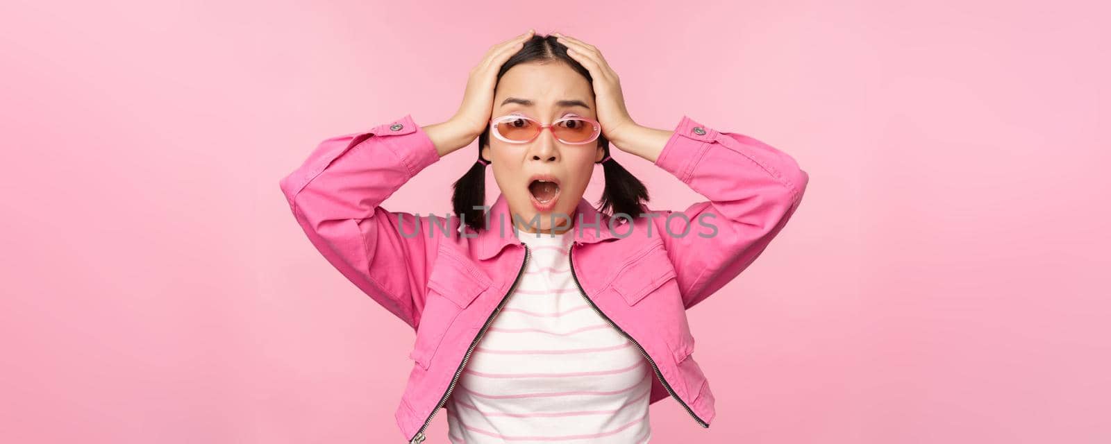 Portrait of troubled asian woman, korean girl holds hands on head and look in panic, standing distressed and shocked against pink background by Benzoix