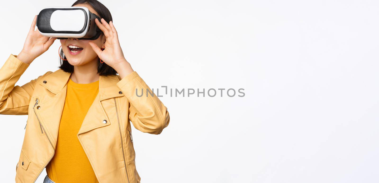 Enthusiastic asian girl in VR glasses, looking with virtual reality headset at smth amazing, standing over white background. Copy space