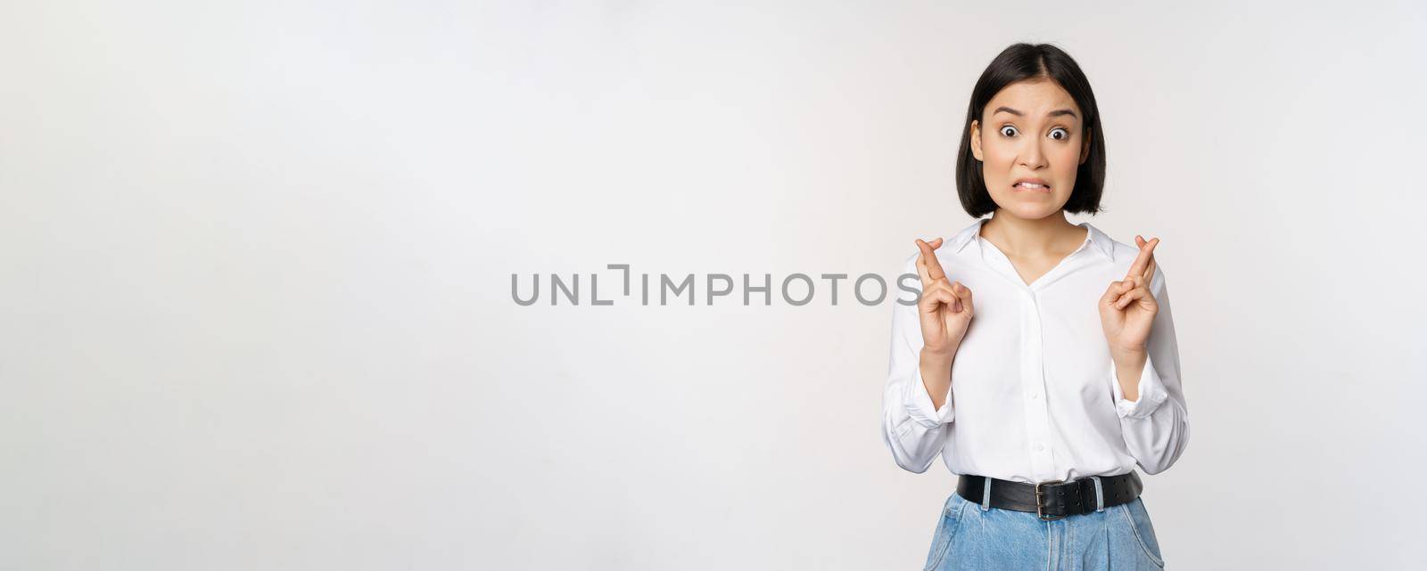 Portrait of young korean woman, asian girl cross fingers and praying, making wish, anticipating, waiting for results, standing over white background by Benzoix