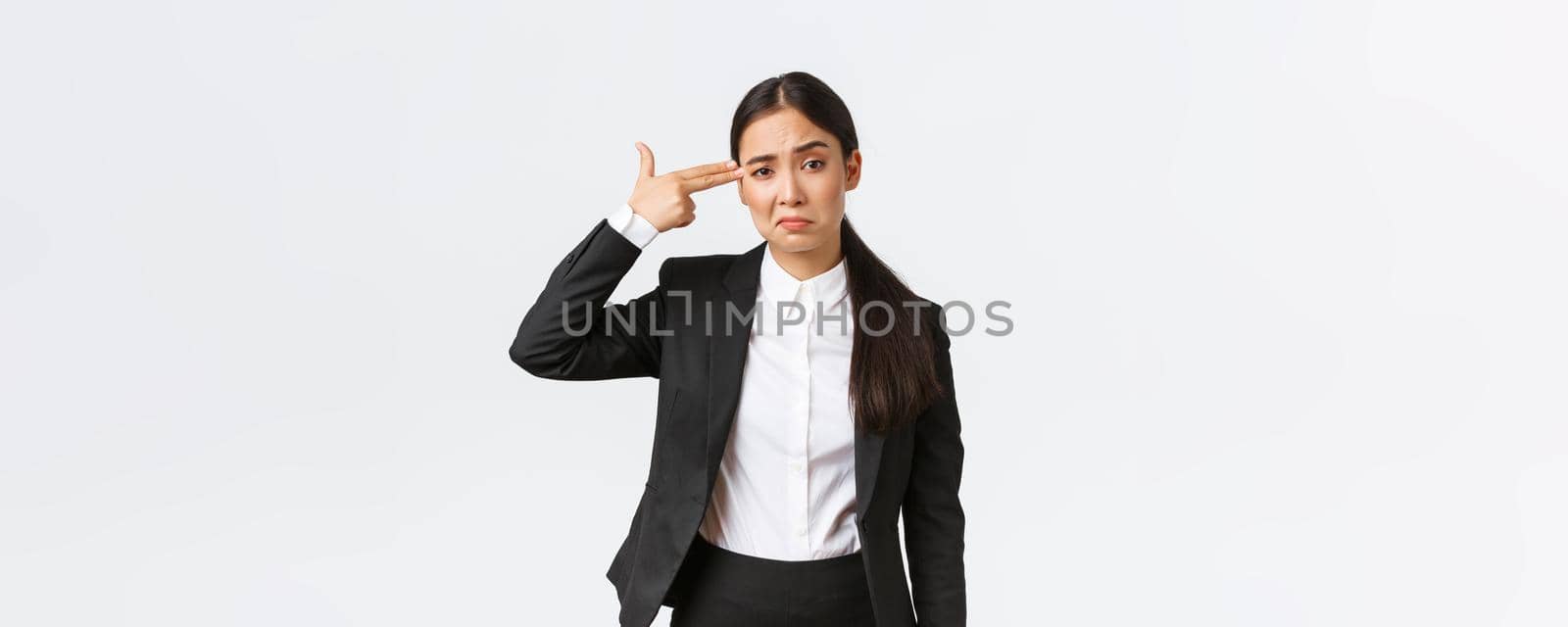 Just shoot me please. Bothered and fed up asian saleswoman hate her work, standing in business suit, shooting herself with hand gun gesture as feeling exhausted, standing white background by Benzoix