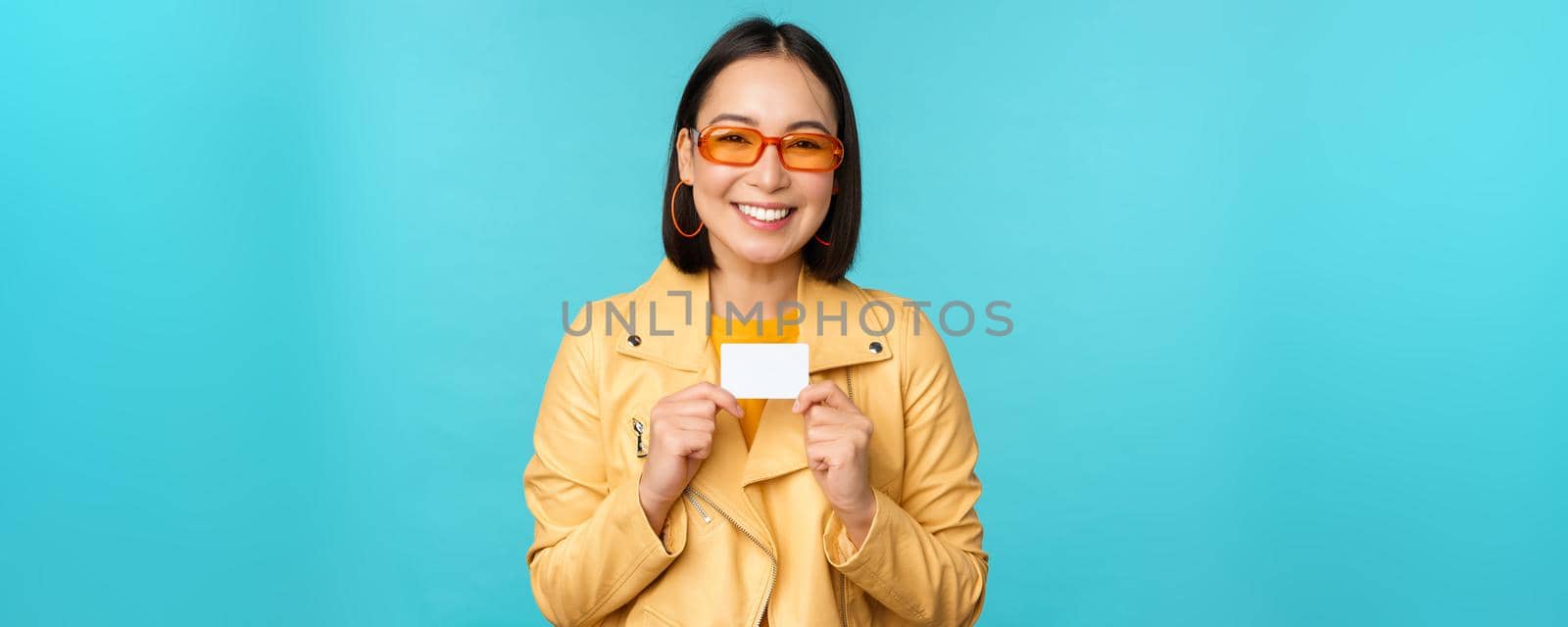 Young beautiful asian woman showing credit card, smiling, choosing bank, standing over blue background by Benzoix