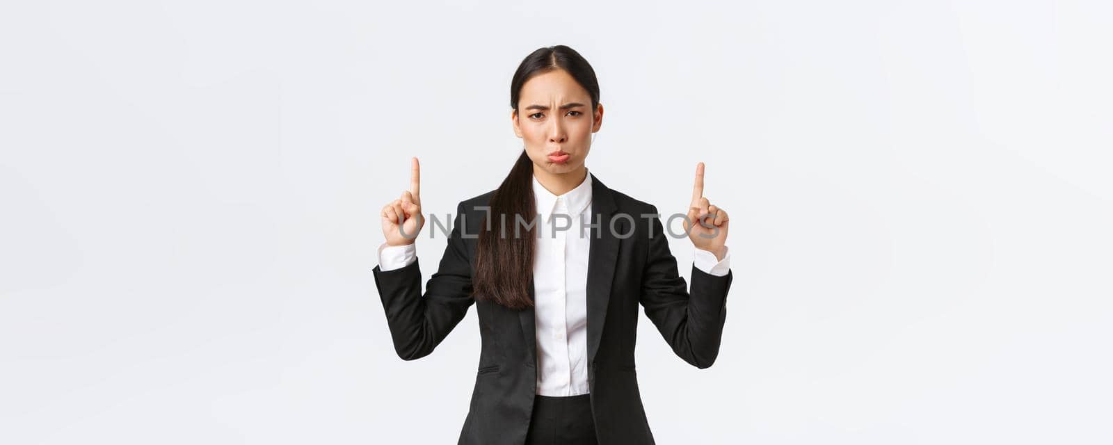 Disappointed pouting asian female entrepreneur in suit complaining and sulking from failure, pointing fingers up, grimacing displeased, feeling down from regret, standing white background.