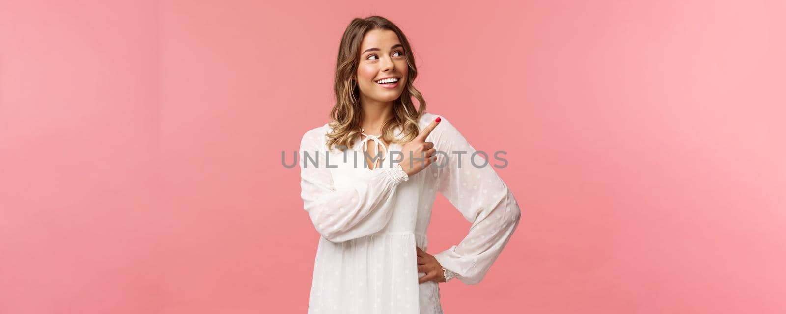 Look at this. Cheerful lovely blond european woman 20s in white cute dress, pointing at upper right corner with beaming smile, looking pleased, satisfied with good offer, pink background.