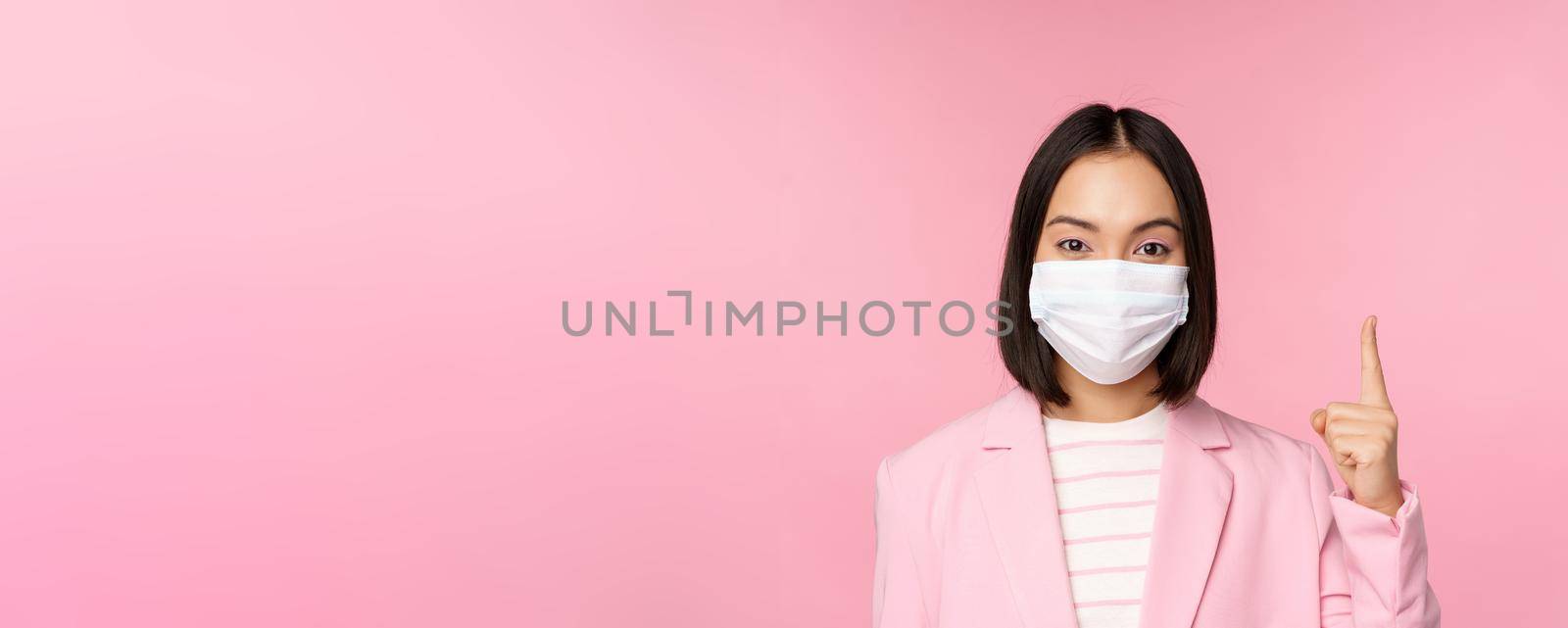 Close up portrait of asian businesswoman in medical face mask and suit, pointing finger up, showing advertisement, top banner, standing over pink background by Benzoix