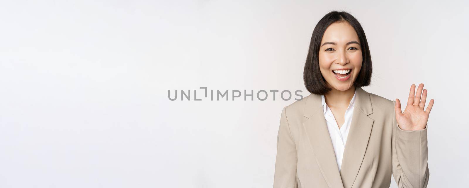 Friendly business woman, asian office lady waving hand and saying hello, hi gesture, standing over white background.