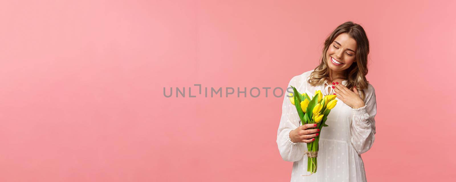 Holidays, beauty and spring concept. Portrait of pleased, cute romantic blond girl receive beautiful bouquet of flowers, looking at yellow tulips and touch heart grateful, smiling.