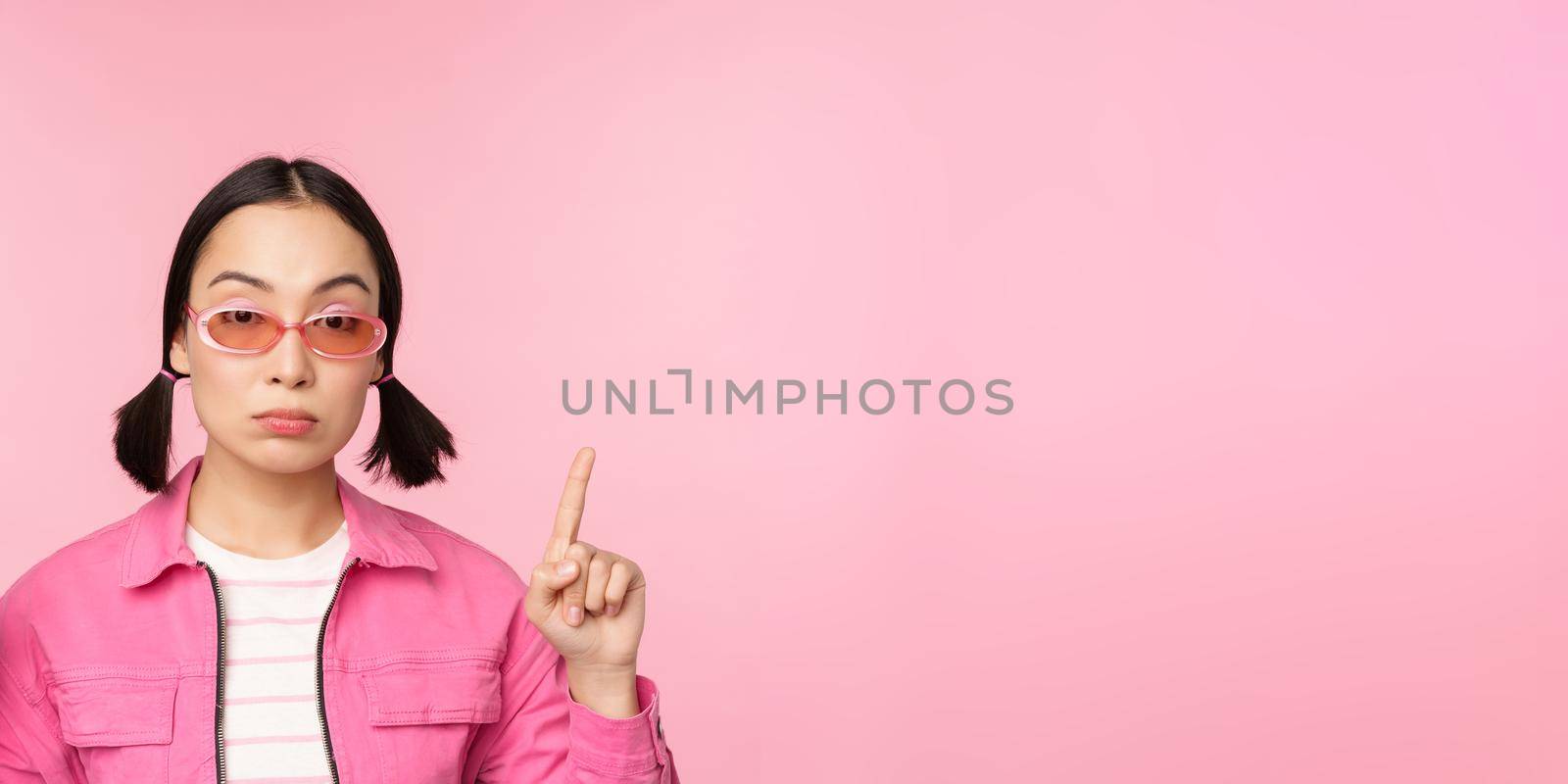 Close up portrait of asian stylish girl, looking skeptical, pointing finger up with unamused face, standing over pink background. Copy space