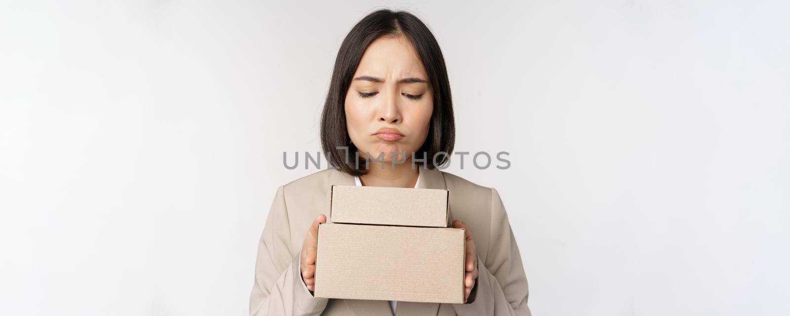 Portrait of asian saleswoman, female entrepreneur holding boxes and looking sad, disappointed, standing over white background.