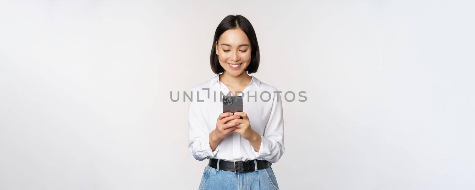 Stylish modern asian girl using mobile phone application, chatting on cellphone and smiling, standing in white blouse against studio background.