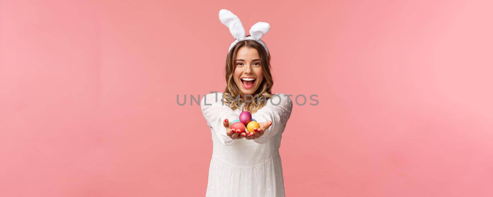 Holidays, spring and party concept. Portrait of lovely, romantic young blond woman in rabbit ears and white dress, giving you painted easter eggs as celebrating orthodox holiday, pink background by Benzoix