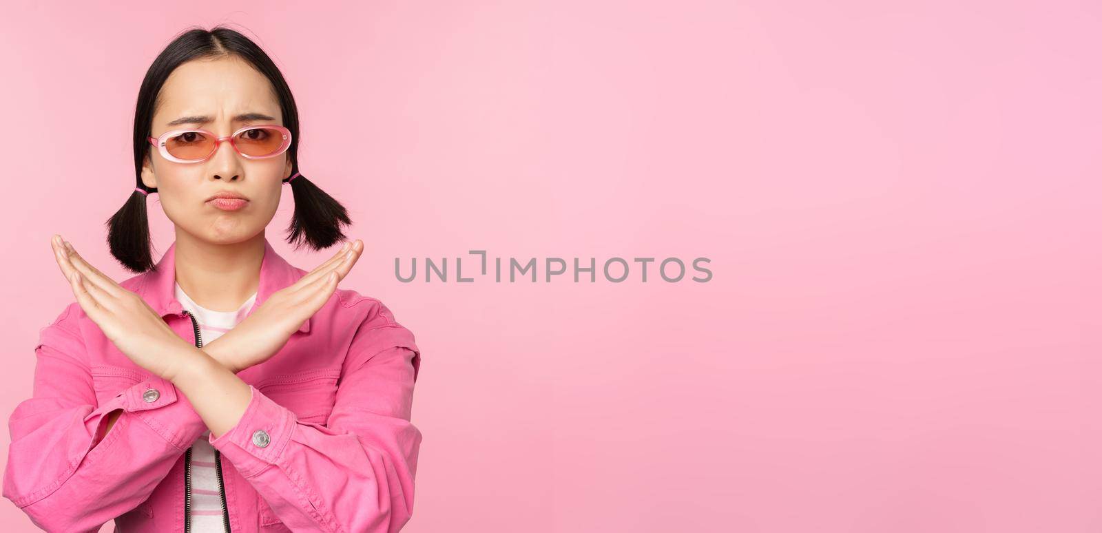 Portrait of korean girl in stylish sunglasses, sulking disappointed, showing stop, rejection gesture, cross sign, standing over pink background by Benzoix