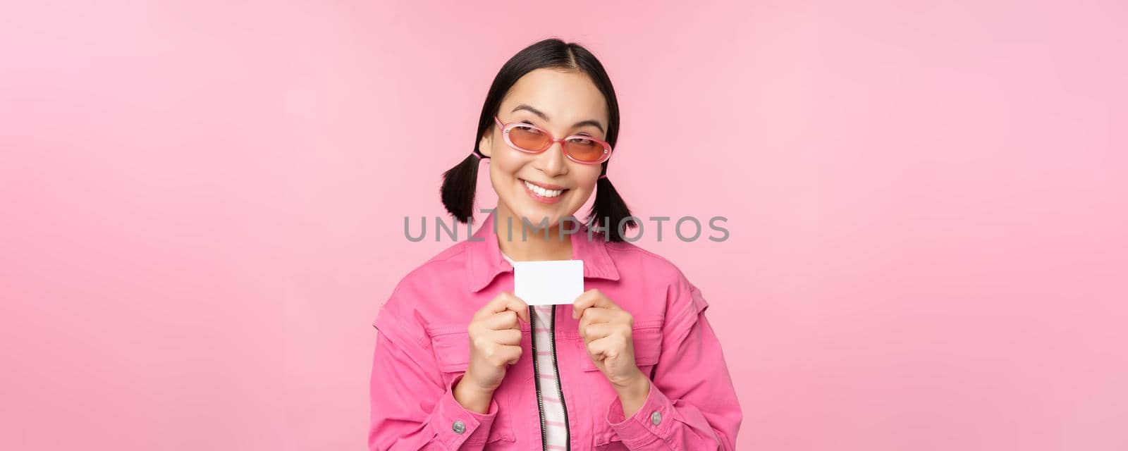 Portrait of stylish, modern asian girl, shows discount, credit card and look pleased, paying contactless, concept of shopping, standing over pink background by Benzoix
